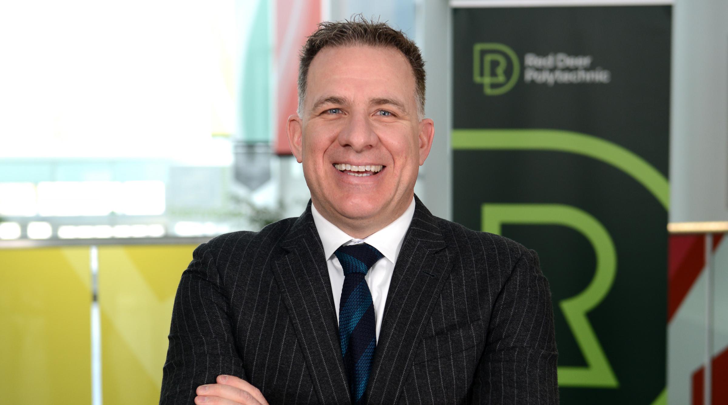 Stuart, wearing a black pinstriped jacket and blue striped tie, laughingly smiles with his arms crossed.