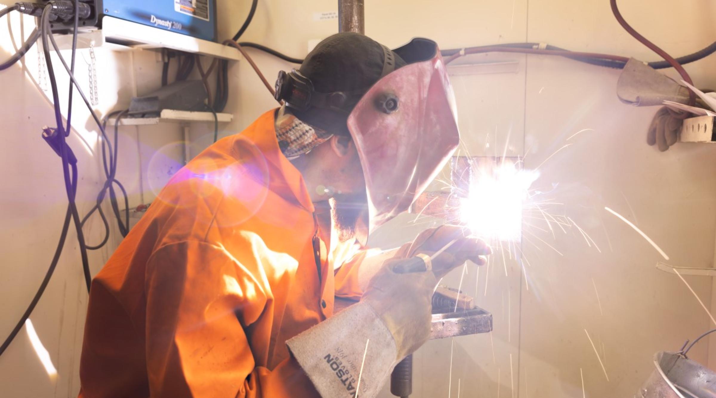 Student in orange coveralls and a welding facemask welds in a shop