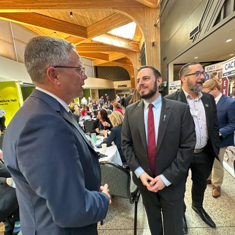 Minister Nicolaides speaks to a gentleman with a large room of people behind him