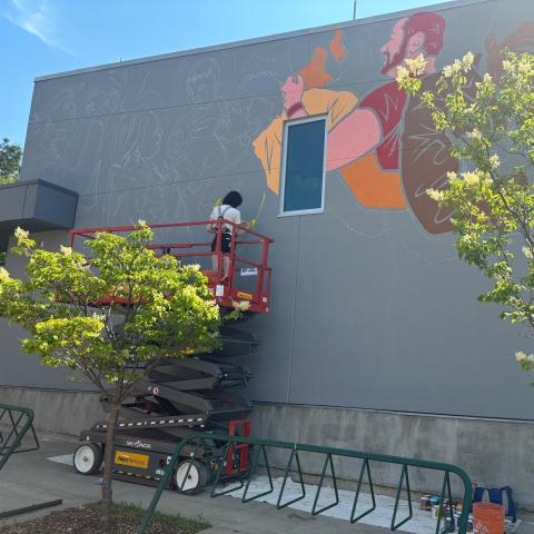 A woman on a scaffold is painting a large vibrant Pride mural on RDP's campus