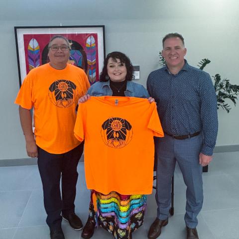 A smiling Cori holds up an orange shirt bearing her design. Dean, Indigenous Iniatives Lloyd Dejarlais and RDP President Stuart Cullum stand on either side of her. 