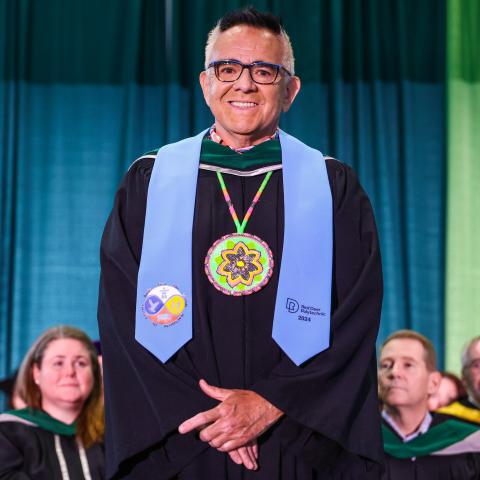 George Littlechild stands on the convocation stage smiling. He wears the light blue stoll with Indigenous imagery and the RDP logo. He also wears a large beaded medallion resembling a flower.