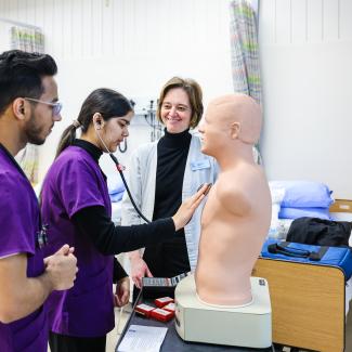 Two Practical Nurse students practice using a stethoscope on a dummy while an instructor watches.
