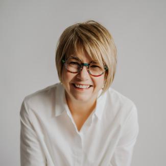 Miji in white shirt, short blonde hair and black glasses sits against a white background.