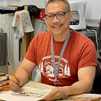 Willie in orange shirt with grey lanyard, works on a painting. the table is covered in paint supplies and other pieces of artwork.