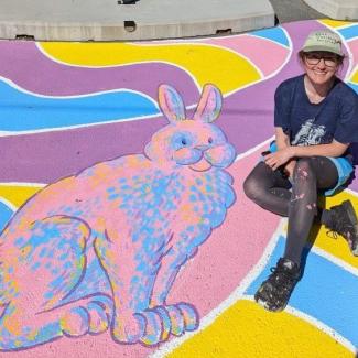 Amber sits on a painting of a brightly colored rabbit on a concrete surface.