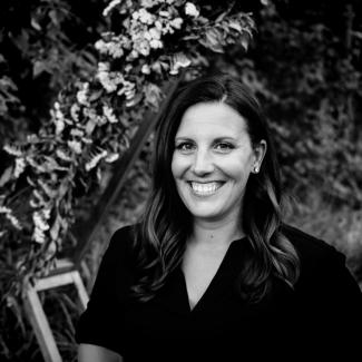 A black and white photo of Becky outside in a dark v neck shirt with long dark hair. She smiles brightly at the camera.