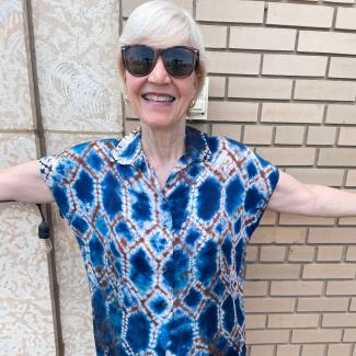 Susan, in a diamond pattern tie dye shirt, smiles brightly with arms outstretched.