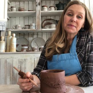 Brenda pauses working on a pottery project. Behind her a white cabinet is full of dry ceramic objects.