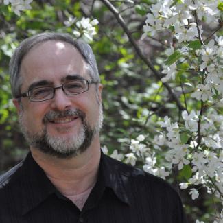 Milt stands under a blossoming apple tree branch. He wears a black shirt and glasses.