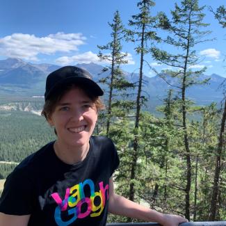 Gillian, wearing a black cap and tshirt, smiles widely in front of a mountain landscape.