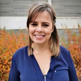 Jana in a blue button up shirt with shoulder length dark blonde hair stands in front of short shrubs with dark orange leaves.