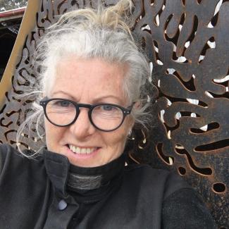 Jen, in black coat  and black rimmed glasses, stands in front of a metal wall with squiggly cut outs.