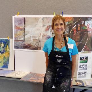 Kathleen, in blue shirt and black apron, stands in front of geometric design paintings.