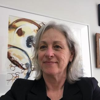 Brenda, in black jacket, sits in front of a large painting with brown and grey swirls.