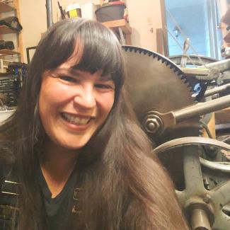 Michelle, with long brown hair, smiles in front of a metal sculpture.