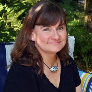 Carol Ann sits outside surrounded by green foliage. She wears a black shirt and necklace with large bead.