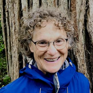 Deborah smiles brightly in a blue jacket standing in front of a large tree. She has short curly light brown hair and glasses.