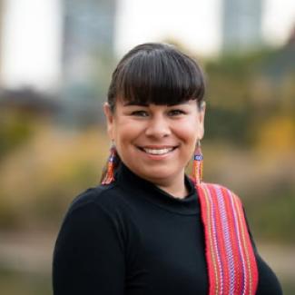 Veronica is wearing a black shirt with a Métis Sash draped over her shoulder. She has a wide smile and dark hair with bangs.