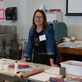 Arlene leans on a table full of art supplies, smiling to the camera.