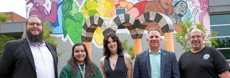Group shot of speakers who presented at the unveiling of new Pride mural. The colourful mural featuring a variety of people in the Pride colours is featured on the wall behind the group. 