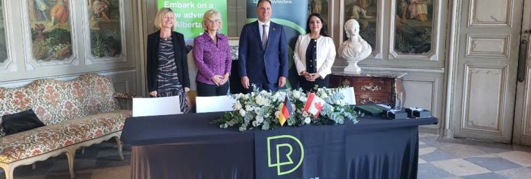 A smiling group of representatives from RDP, EUF and HSBI stand in front of a table with Canadian and German flags 