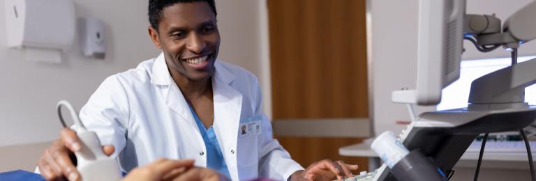 A sonographer smiles while using an ultrasound wand on a patient abdomen.