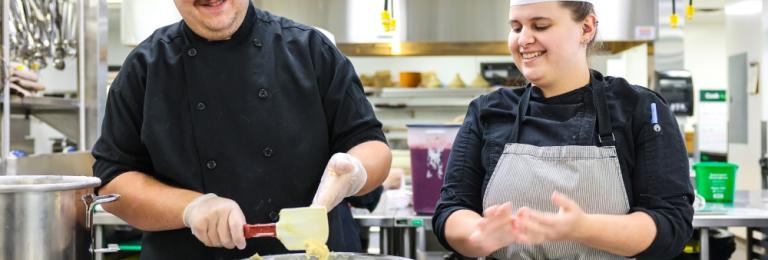 A smiling male and female student in chef's hats prepare some baking in a commercial kitchen