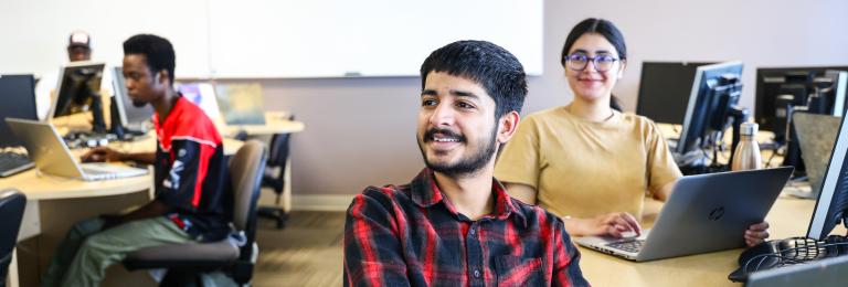 A, student in a plaid shirt, smiles to the instructor (who is not seen) in a computer lab classroom.