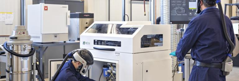 Two men wearing blue lab coats and protective helmets are working with the ExOne 3D Printer