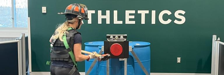 A female in a firefighter helmet and harness is swinging a hammer against a large red target as part of the Firefighter Applicant Physical Test