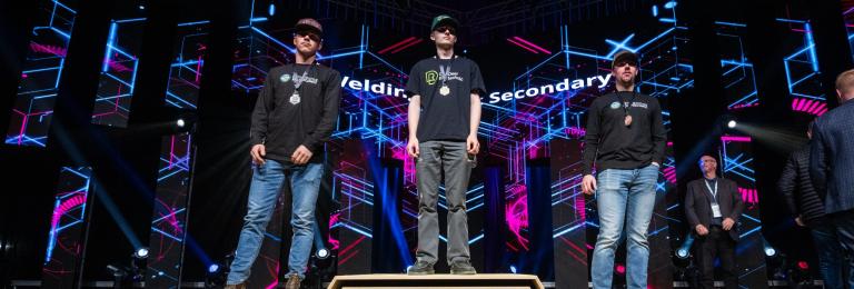 Lucas Stang (RDP Welding student) stands with his gold medal on a podium with two other welders at Skills Canada Alberta 2025