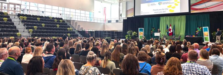 A large crowd sits in the GW Harris main gym looking towards a keynote speaker who's standing on the main stage