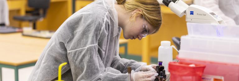 A female student in protective gear works with small bottles of chemical compounds in a lab