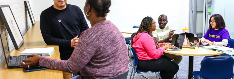 A man chats with a Black woman who has her back turned to the camera. In the background, a group of Black students chats at a round table