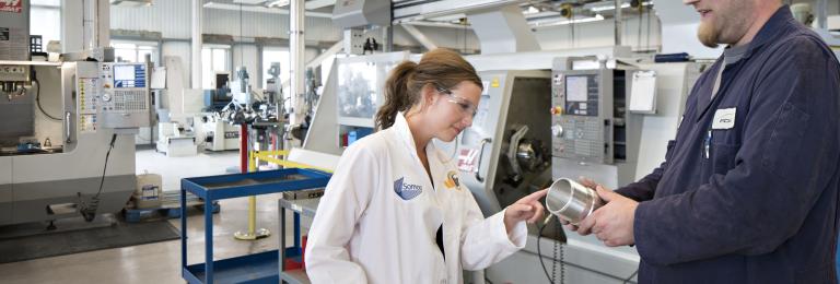 A female student in a lab coat examines a part being shown to her by a male lab tech in the CIM-TAC