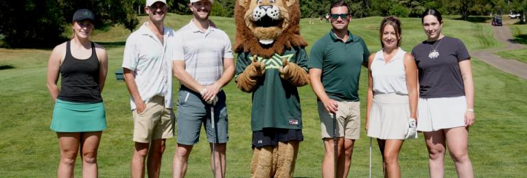 A group of golfers, student athletes, and Rufus the lion stand on a golf course fairway