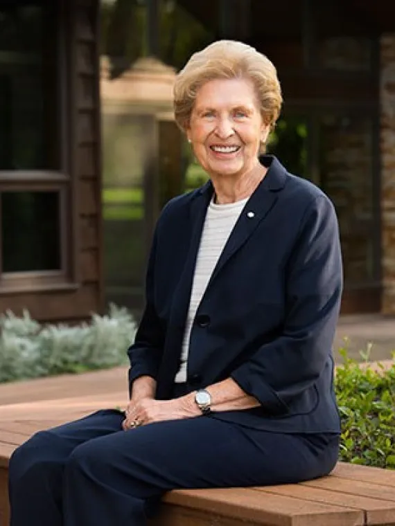 Joan Donald smiles while sitting on a wooden bench in a dark blue pant suit in front of a house