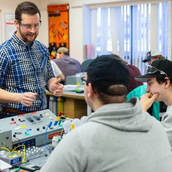 An electrician faculty member speaks to two students who are working with a control panel of wires and buttons.