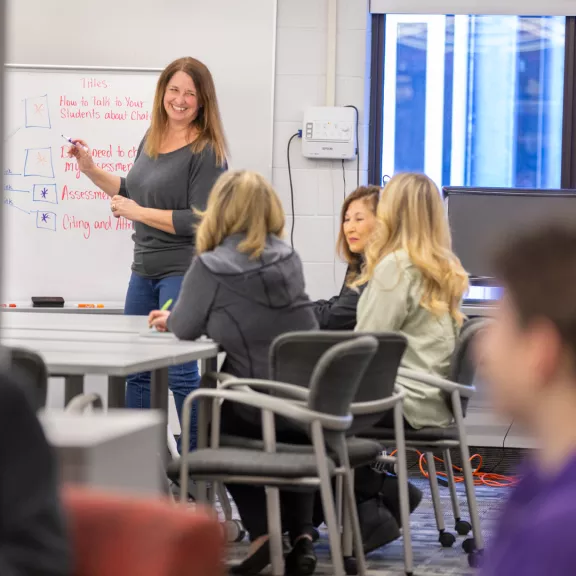 An instructor smiles at a class of students while standing in front of a whiteboard.