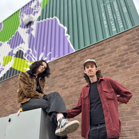 Two individuals stand in front of a brick wall with a brightly coloured mural while looking down at the camera