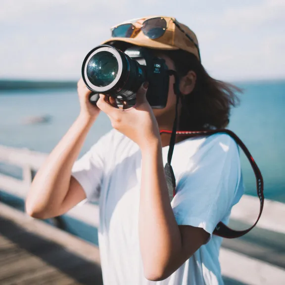 Someone in a hat holds a large camera in front of their faces while standing on a pier