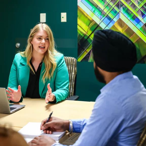 A woman in a green blazer sits at the head of a table speaking to a person whose back is to the camera.