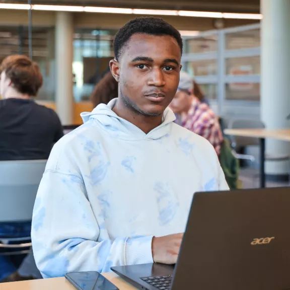 A Black male student sits in front of a laptop in a library looking at the camera