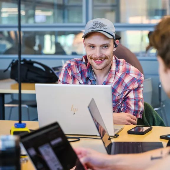 A student in a plaid shirt and grey cap smiles at a fellow student. They have laptops & phones on the table in front of them.