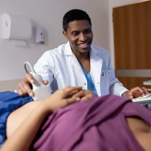 A sonographer smiles while using an ultrasound wand on a patient abdomen.