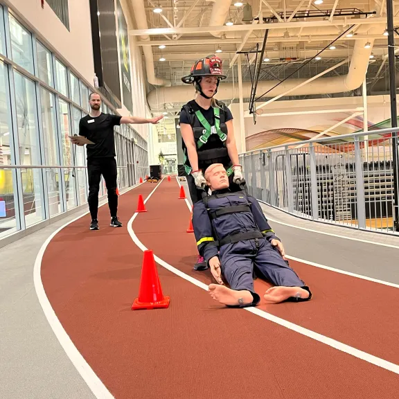 A woman in weighted vest carries a weighted manakin through a course as a personal trainer directs her