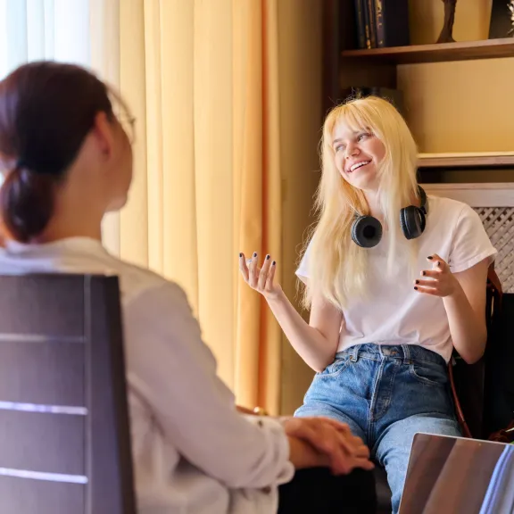 A smiling, energetic young lady talks while sitting across from a woman who has her back to the camera