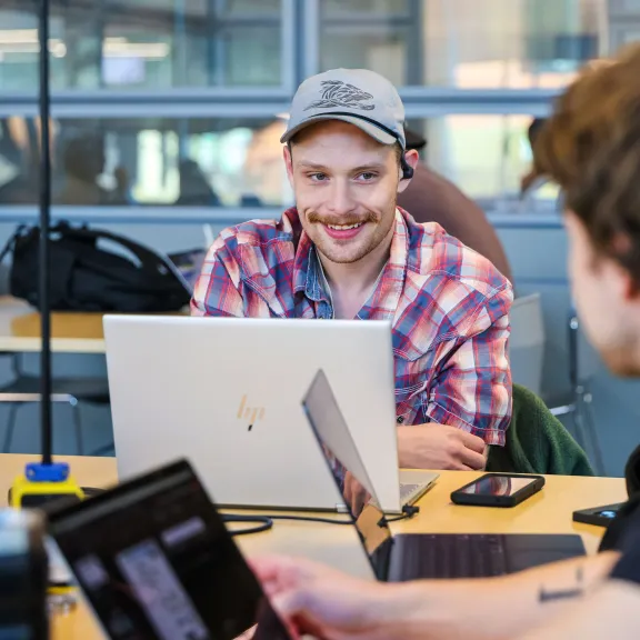 A student in a plaid shirt and grey cap smiles at a fellow student. They have laptops & phones on the table in front of them.