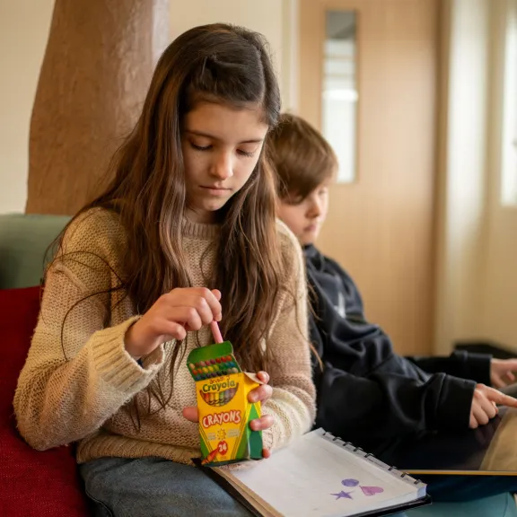 A young girl is picking out crayons from a box with a picture on her lap. A boy sits behind her